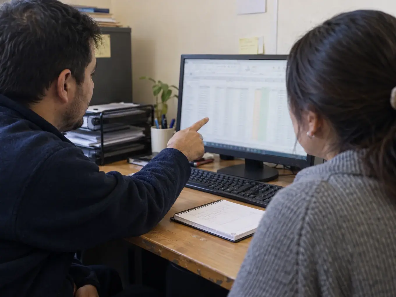 Un asesor señala una planilla en el monitor mientras una clienta la revisa en su escritorio
